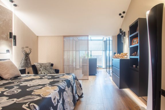 Habitación moderna con cama, estantería y luz natural entrando por la ventana.