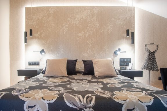 Habitación con cama moderna, luces ambientales en cabecero y rodapié, alicatado con pasta blanca imitación yute de Porcelanosa.